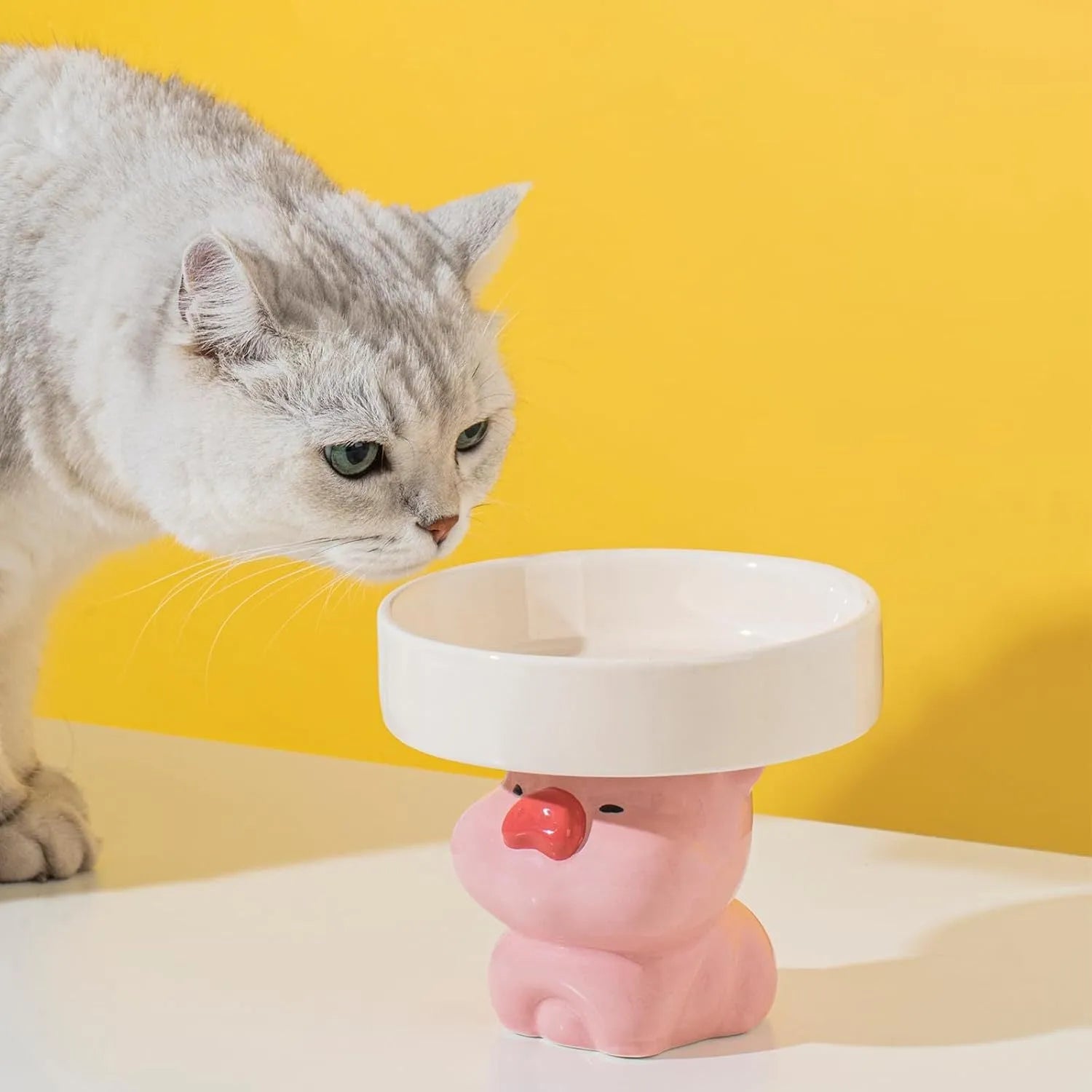 Raised ceramic cat bowl with animal design in pink, yellow, or white, showing wide feeding surface and elevated base.