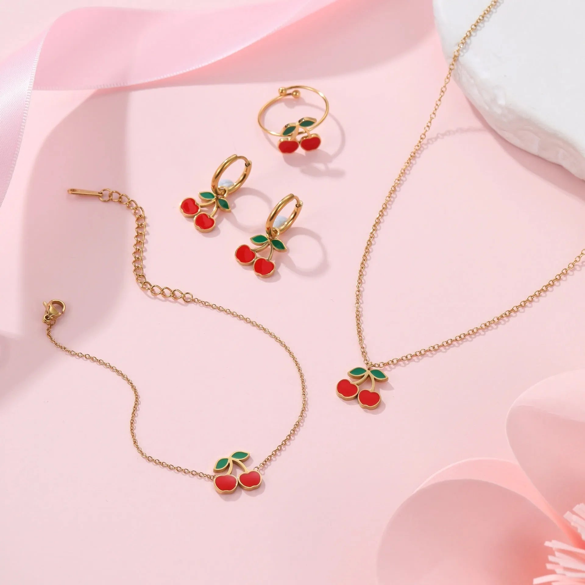 Four-piece cherry-themed jewellery set with gold electroplated necklace, bracelet, earrings, and ring on a white background.