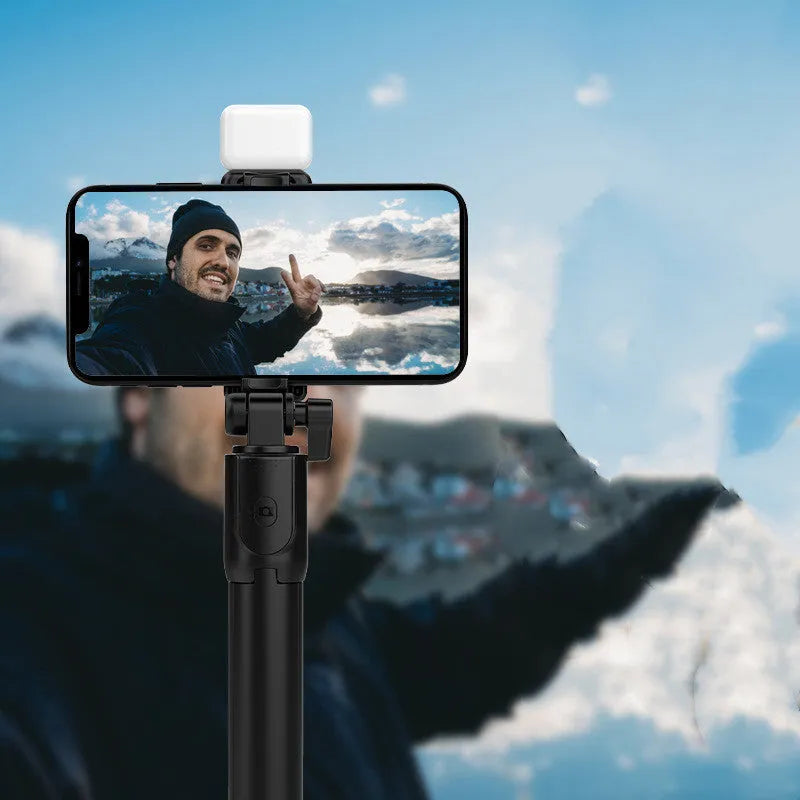 Black Bluetooth selfie stick with extended tripod legs and a fill light, holding an iPhone.