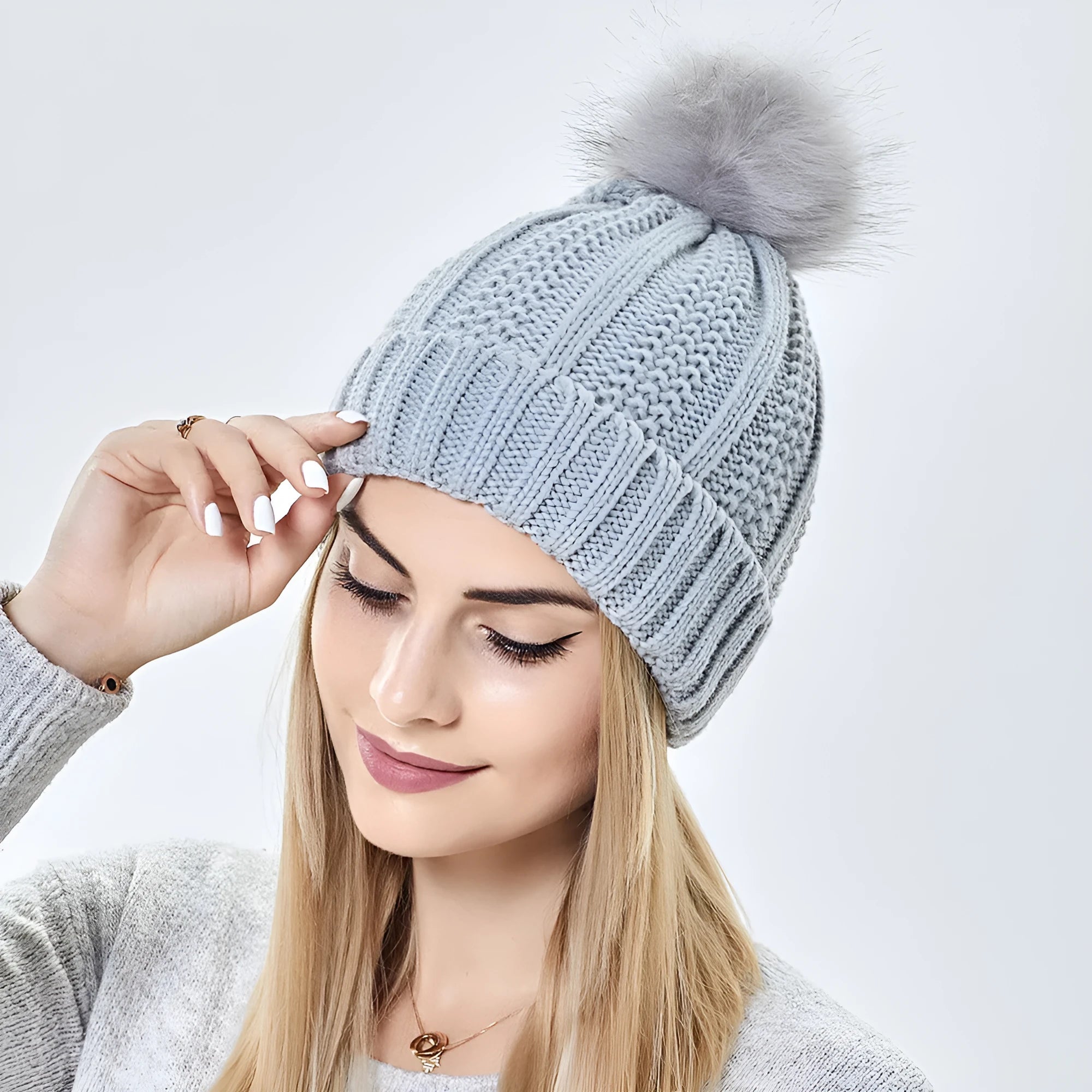 Woman wearing a black knitted beanie with a grey faux fur pom pom, showing the satin lining.