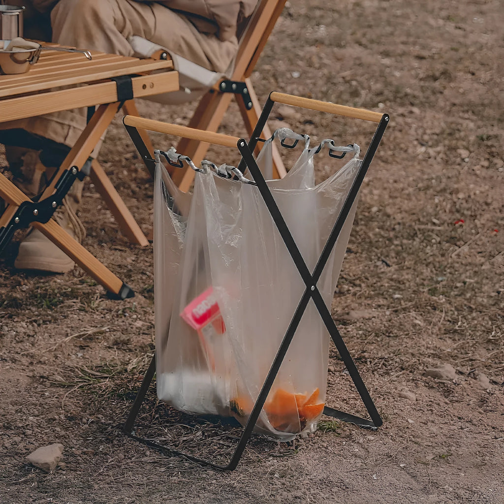 Black folding iron trash rack with a wooden handle holding a rubbish bag outdoors.