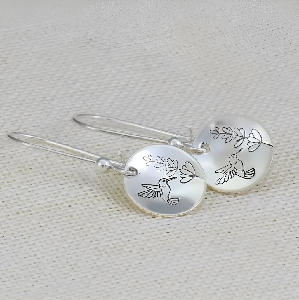 Silver-coloured round drop earrings with a detailed bird and floral design against leaves.
