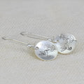 Silver-coloured round drop earrings with a detailed bird and floral design against leaves.