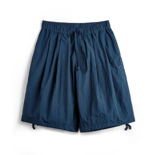 Men's loose Prussian Blue nylon shorts with embroidery on a beach background.