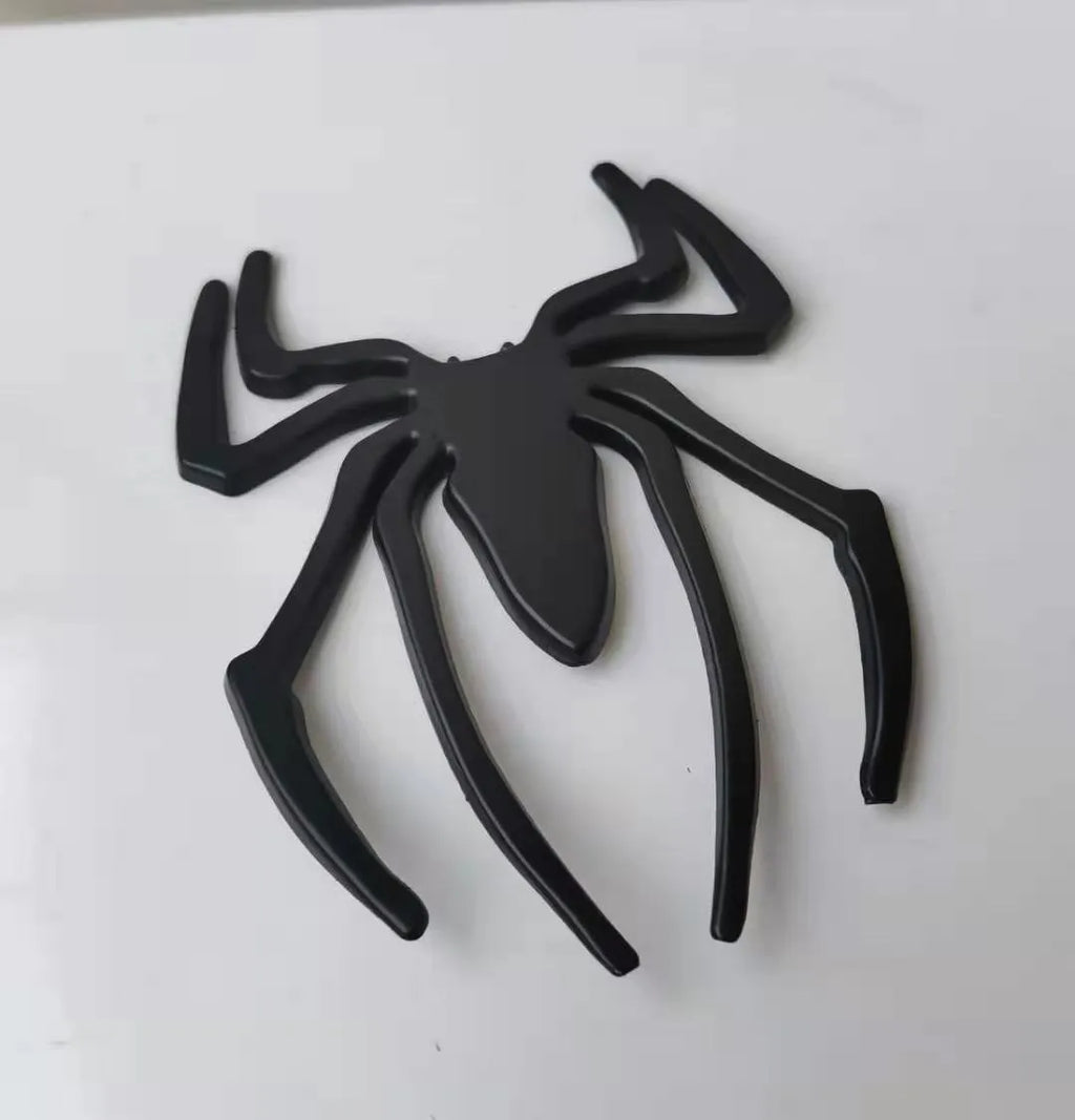 Three-dimensional metal spider bumper sticker in metallic finish on a car bumper.