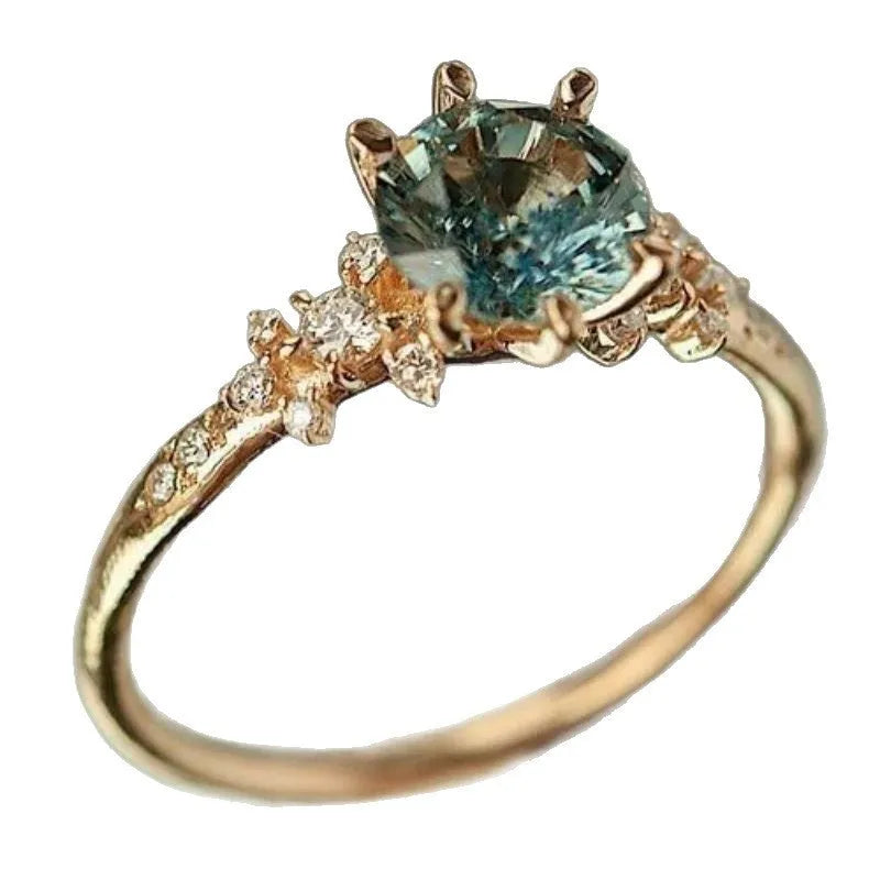 A gold-coloured geometric ring with a claw-set zircon stone and a chamfered square pattern on the band.